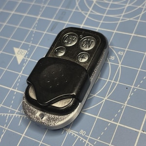 May include: A black and silver remote control with "OFF" and "ON" buttons. The remote has a black cover and plus and minus buttons. The remote is on a blue cutting mat with white grid lines and numbers.