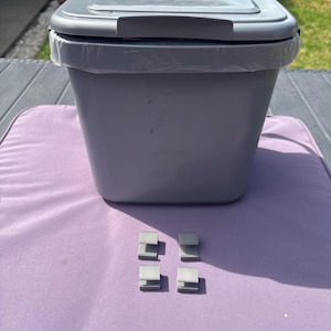以下が含まれることがあります： ヒンジ付きの蓋が付いたライトグレーのプラスチック製ゴミ箱で、縁の周りに透明なプラスチックライナーが見えます。ゴミ箱の前には、金属色の小さな正方形のクリップ4つが2列2行のグリッド状に配置されています。