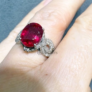 Può includere: Un anello d'argento con una grande pietra preziosa rosso scuro a taglio ovale. La gemma è il punto focale, incastonata in una fascia d'argento dettagliata e ornata con pietre più piccole. L'anello è mostrato su un dito.