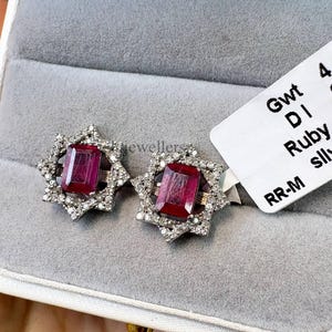 May include: A pair of silver stud earrings with a starburst design. Each earring features a rectangular, deep red ruby stone surrounded by small, clear diamonds. The earrings are displayed in a grey jewellery box.
