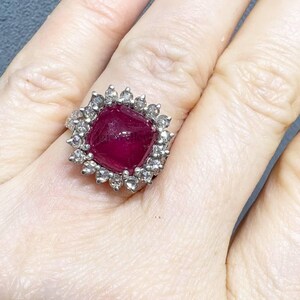 May include: A square-cut, deep red gemstone ring with a halo of smaller, clear stones. The ring is set in a silver-colored metal and is worn on a finger. The ring's design is classic and elegant, suitable for special occasions.