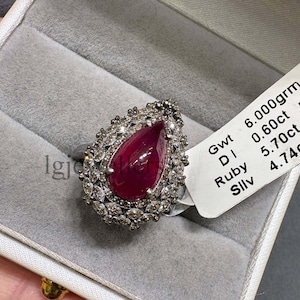 May include: A pear-shaped ruby ring surrounded by a halo of small white stones, presented in a white jewellery box. A white tag attached to the ring displays the text: Gwt 6.000grm, DI 0.60ct, Ruby 5.70ct, Sllv 4.74grm, and the code RR-M.