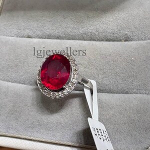 May include: A silver ring featuring a large, oval, deep red gemstone encircled by a halo of small, clear stones. The ring is presented in a white jewellery box with a grey interior. The text "lgjewellers" is visible.