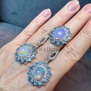 May include: A jewelry set consisting of a ring and two drop earrings, each featuring a central oval cabochon opal surrounded by a halo of small, square-cut light blue gemstones and a secondary border of tiny clear white stones in silver settings.