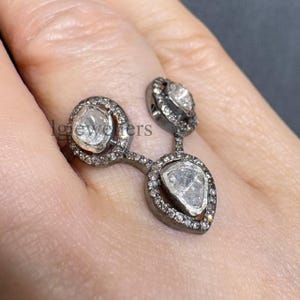 Peut inclure: Une bague en métal sombre ornée de trois pierres blanches translucides et irrégulières disposées en grappe. Les pierres sont entourées de petites pierres d'accent blanches serties pavé. Le texte lgjewellers est visible sur la bague.