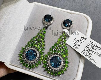 May include: A pair of silver earrings with a large, round, dark blue gemstone surrounded by small white stones. The earrings have a teardrop shape, with green gemstones fanning out from the center. The earrings are displayed in a white jewelry box.