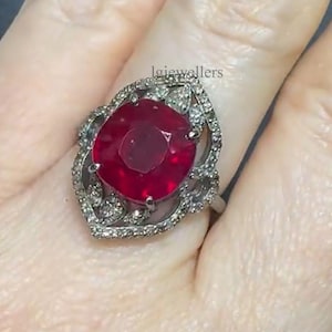 Puede incluir: Un anillo de plata con una gran gema roja oscura tallada en cojín. La gema está rodeada de piedras más pequeñas y transparentes y un engaste ornamentado de filigrana. El anillo se muestra en un dedo.