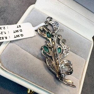 May include: A silver-toned brooch with an ornate floral design, featuring oval-cut green gemstones and small clear stones. The brooch is displayed in a white jewelry box with a gray interior. A white tag with text is attached.