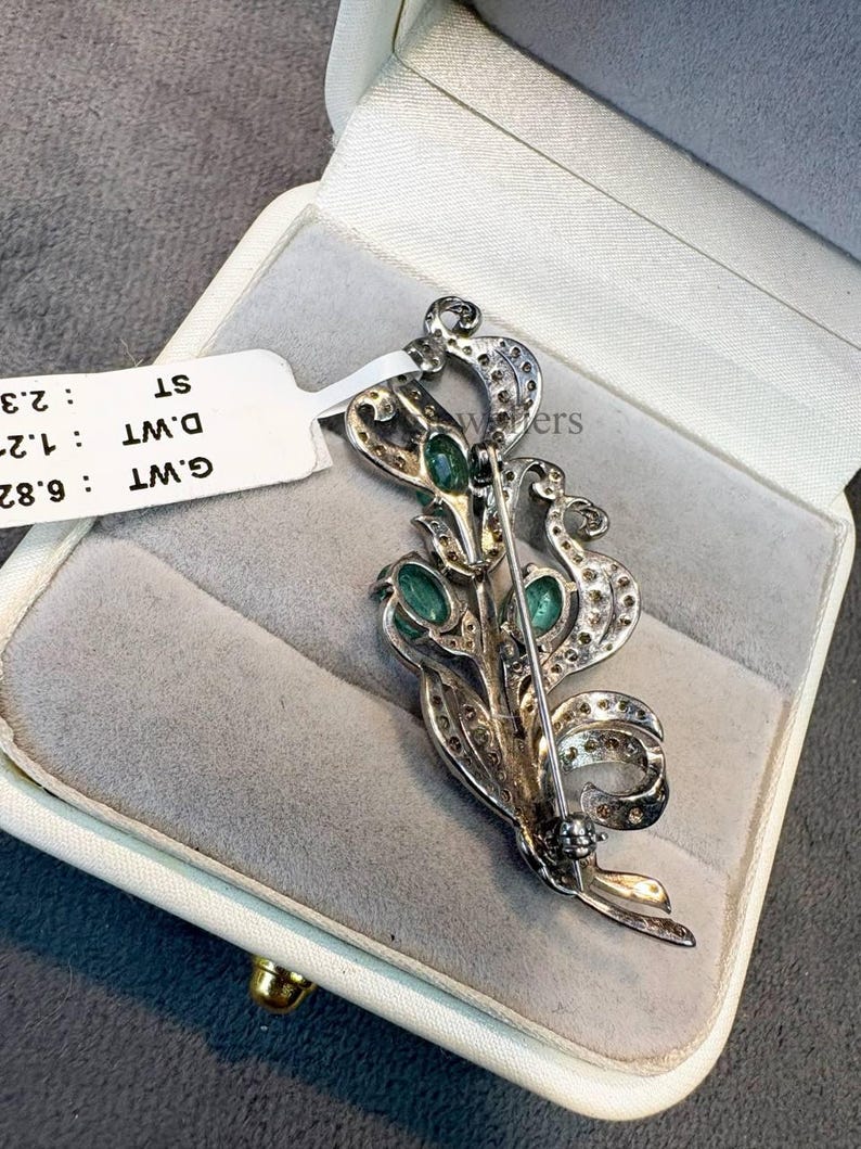 May include: A silver-toned brooch with an ornate floral design, featuring oval-cut green gemstones and small clear stones. The brooch is displayed in a white jewelry box with a gray interior. A white tag with text is attached.