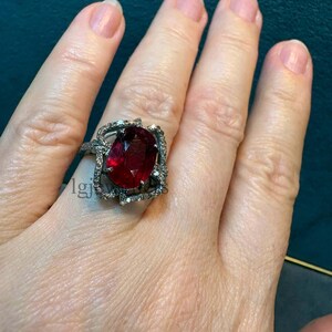 Op de afbeelding: Een ring met een grote, ovaal geslepen dieprode edelsteen in een donker metalen zetting. De zetting is omgeven door een decoratieve, opengewerkte rand die is ingelegd met kleine, heldere, fonkelende steentjes.