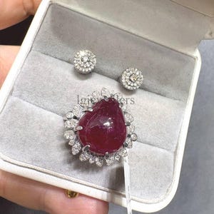 Op de afbeelding: Een sieradenset bestaande uit een ring met een grote, peervormige, dieprode robijn omringd door een halo van kleine, heldere, ronde diamanten, vergezeld door twee bijpassende ronde oorknopjes met een centrale steen en kleine diamanten.