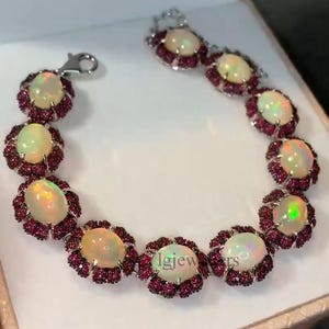 Peut inclure: Un bracelet en argent orné d'opales ovales et de petites pierres rouges foncées rondes. Les opales ont un aspect laiteux et irisé. Le bracelet est composé de maillons en forme de fleurs, reliés par un fermoir. Longueur approximative : 18 cm.