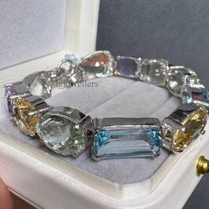 Può includere: Un bracciale color argento caratterizzato da una serie di pietre preziose sfaccettate in varie forme, tra cui tagli ovali, rettangolari e a goccia. Le pietre presentano sfumature azzurre, gialle, viola e trasparenti.