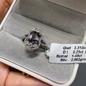 Puede incluir: Un anillo de plata con una piedra central rectangular de espinela gris oscuro rodeada por un halo de pequeños diamantes blancos en un engaste decorativo. Una etiqueta blanca unida al anillo muestra el texto: Gwt 3.310ct, DI 0.25ct, Spinal 1.49ct, Sllv 2.962grm.