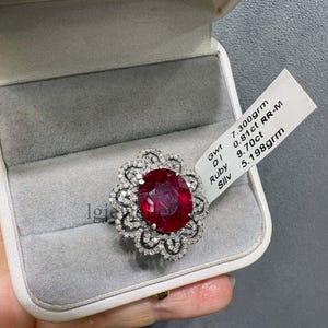 Puede incluir: Un anillo con un gran rubí rojo de corte ovalado rodeado por un halo floral de pequeños diamantes blancos. Una etiqueta blanca unida al anillo muestra el texto: Gwt 7.300grm, DI 0.81ct RR-M, Ruby 9.70ct, Sllv 5.198grm.