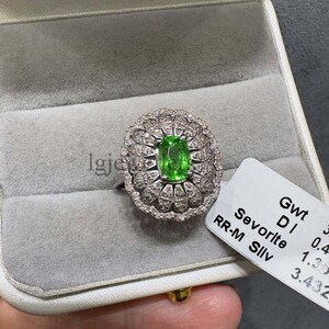 Puede incluir: Un anillo de plata con una tsavorita verde de corte ovalado en el centro, rodeada por un halo festoneado de pequeños diamantes redondos. Una etiqueta blanca unida al anillo muestra el texto Gwt, DI 0.4, Sevorite 1.35 y RR-M Silv 3.432.