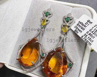 May include: A pair of elegant drop earrings featuring large, teardrop-shaped orange gemstones. The earrings have a silver-colored setting with smaller green and yellow stones, and are displayed in a white jewelry box. The earrings are labeled "lgjewellers".