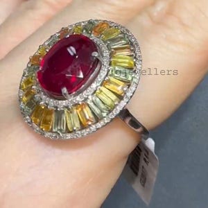 Puede incluir: Anillo ovalado con una piedra roja oscura en el centro, rodeada por un halo de piedras amarillas y verdes. El engaste está adornado con pequeñas piedras claras. El anillo se muestra en un dedo.