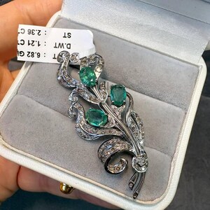 May include: A silver-toned brooch in a floral design, featuring three oval emeralds and numerous small diamonds. The brooch is presented in a white jewelry box with a gray interior. The brooch is a piece of fine jewelry.