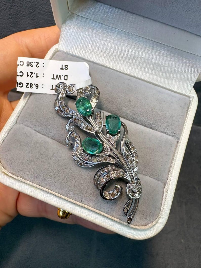 May include: A silver-toned brooch in a floral design, featuring three oval emeralds and numerous small diamonds. The brooch is presented in a white jewelry box with a gray interior. The brooch is a piece of fine jewelry.