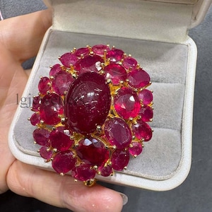 May include: A large gold ring featuring a prominent central oval-cut deep red gemstone surrounded by a cluster of smaller oval-cut red gemstones. The ring is set in a yellow gold metal frame and is presented inside a light grey velvet-lined jewellery box.