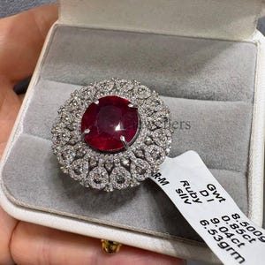 Könnte beinhalten: Ein silberfarbener Ring mit einem großen, oval geschliffenen, tiefroten Rubin, umgeben von einem Kranz aus kleinen, rund geschliffenen weißen Steinen. Ein weißes Etikett am Ring zeigt den Text: Gwt 8.500g, D 0.85ct, Ruby 9.04ct, sllv 6.53grm.