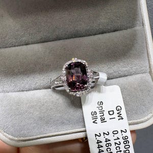 Op de afbeelding: Een zilveren ring met een kussengeslepen paarse spinel edelsteen omringd door een halo van kleine witte diamanten. Een wit label aan de ringband toont de tekst: Gwt 2.960grm, D 0.12ct RR-M, Spinal 2.46ct, Silv 2.444grm.