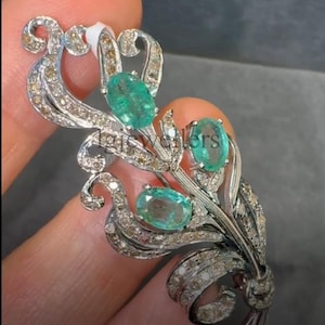 May include: A silver-toned brooch with an ornate floral design. The brooch features three oval-cut, bright green gemstones and is embellished with small, sparkling diamonds. The design includes curved lines and a bow at the base, creating an elegant accessory.
