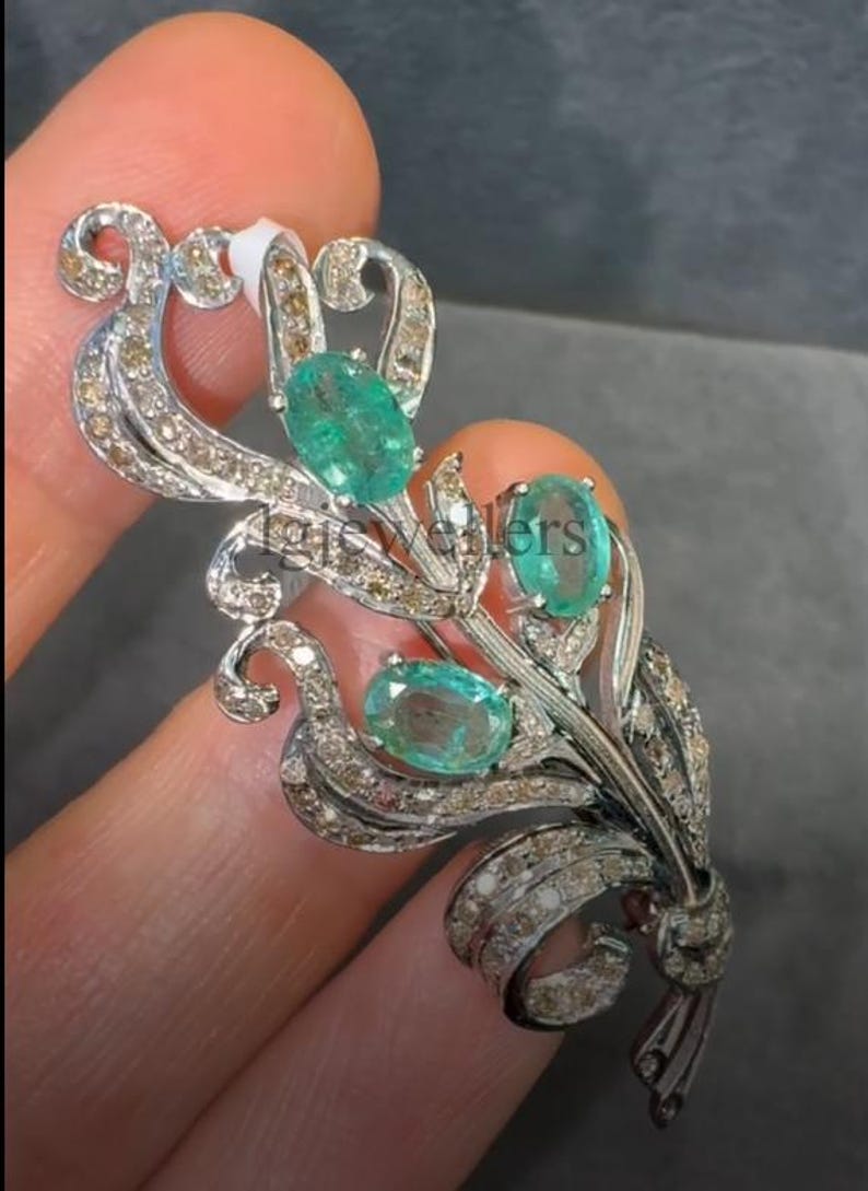 May include: A silver-toned brooch with an ornate floral design. The brooch features three oval-cut, bright green gemstones and is embellished with small, sparkling diamonds. The design includes curved lines and a bow at the base, creating an elegant accessory.