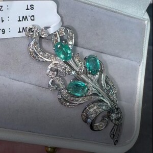 May include: A silver-toned brooch featuring three oval, teal-colored gemstones. The brooch has a floral design with a ribbon detail and is encrusted with small, clear gemstones. The brooch is displayed in a gray jewelry box.