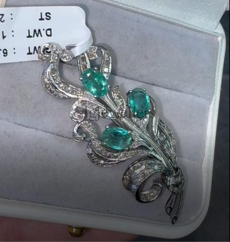 May include: A silver-toned brooch featuring three oval, teal-colored gemstones. The brooch has a floral design with a ribbon detail and is encrusted with small, clear gemstones. The brooch is displayed in a gray jewelry box.