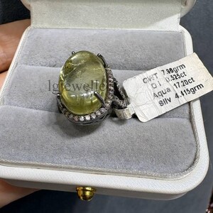 Puede incluir: Un anillo con una gran piedra preciosa ovalada de color amarillo pálido engastada en una banda de metal oscuro rodeada de pequeñas piedras blancas. Una etiqueta blanca muestra el texto: GWT 7.98grm, DI 0.525ct, Aqua 17.20ct, Sllv 4.415grm. El anillo está en una caja gris.