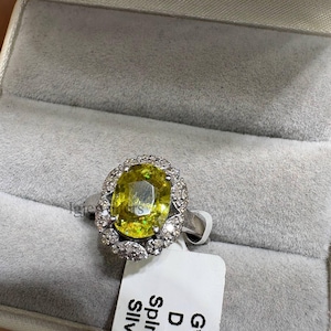 Puede incluir: Un anillo de plata con una gran piedra preciosa de color amarillo verdoso y corte ovalado, rodeada por un halo de pequeñas piedras claras de corte redondo. Una etiqueta blanca adjunta muestra el texto Gwt 4, DI 0., Spin 3.4 y Sllv 3.3.