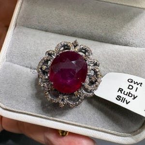 May include: A ring featuring a large, oval-cut, deep red ruby centre stone surrounded by a floral-shaped halo of small, dark-set accent stones. A white tag attached to the ring displays the text Gwt, DI, Ruby, and Silv in black lettering.