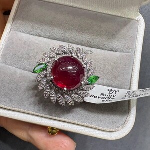 Könnte beinhalten: Ein Ring mit einem großen, runden, tiefroten Rubin im Cabochon-Schliff, umgeben von einem Kranz aus kleinen, klaren, im Pavé-Stil gefassten Diamanten in Blattform, akzentuiert durch zwei grüne Tsavorite im Marquise-Schliff an den Seiten.