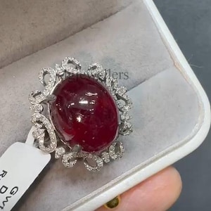 May include: A silver-toned ring featuring a large, oval-shaped, deep red cabochon gemstone at its center. The setting is surrounded by a decorative, openwork metal frame encrusted with small, clear, sparkling stones. A white tag is attached to the side.