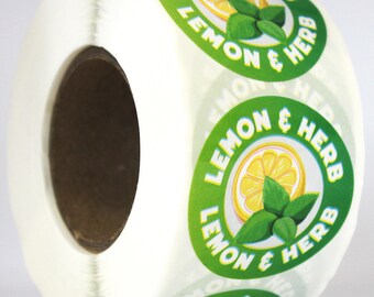 Puede incluir: Un rollo de etiquetas adhesivas circulares con fondo verde, una rodaja de limón amarilla y hojas de hierbas verdes. El texto LEMON & HERB está impreso en letras mayúsculas blancas alrededor del borde circular de cada pegatina del rollo.