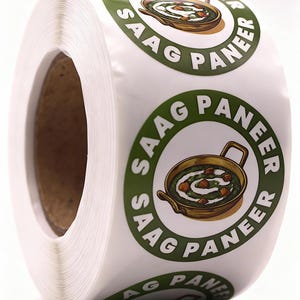 May include: A roll of white stickers with a green and brown circular design. The design features the text "SAAG PANEER" around an illustration of a dish in a pan. The dish appears to be a curry with paneer cheese.