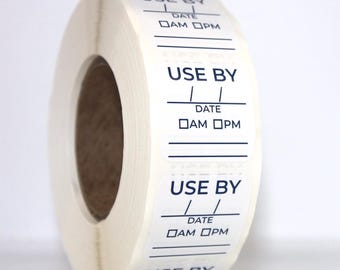 Puede incluir: Un rollo de etiquetas adhesivas blancas con texto impreso en azul oscuro. Cada etiqueta rectangular incluye las palabras USE BY, una línea para la fecha, casillas para AM y PM, y líneas en blanco adicionales para escribir información.