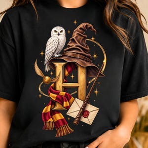 May include: A black t-shirt featuring a graphic print of a large gold letter H, a brown pointed wizard hat, a white owl, a red and yellow striped scarf, a golden winged ball, a wooden magic wand, and a white envelope with a red wax seal.