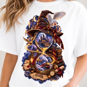 May include: A white t-shirt featuring a graphic print of a brown wizard hat atop an hourglass. Inside the glass are a castle and a steam train. Surrounding elements include a white owl, stacked books, a golden snitch, envelopes, and a pocket watch.