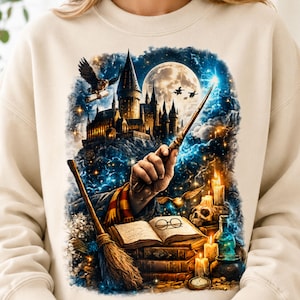 May include: A beige crewneck sweatshirt featuring a graphic print of a castle under a full moon, a hand holding a wand, an open book with glasses, a broomstick, a skull, candles, and a flying owl against a dark blue starry background.