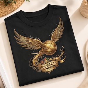 May include: A black t-shirt featuring a gold-coloured winged ball graphic with intricate patterns. Below the ball is a banner with the text HOGWARTS QUIDDITCH and four small house crests. The design includes swirling gold filigree accents.