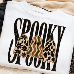 May include: A white crewneck sweatshirt featuring the word SPOOKY in large, distressed black letters. Overlaid on the text are three ghost-shaped patches with stitched edges, featuring floral, wavy striped, and leopard print patterns in neutral tones.