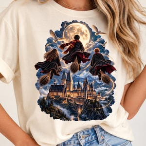 May include: A cream-coloured t-shirt featuring a graphic print of three figures in dark robes flying on broomsticks above a large castle at night. A full moon shines in the background, surrounded by clouds and flying owls, with light trails.