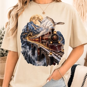 May include: A beige short-sleeve t-shirt featuring a circular graphic print of a red steam locomotive crossing a stone bridge towards a castle in a snowy mountain landscape, with owls flying in the sky and a large full moon in the background.