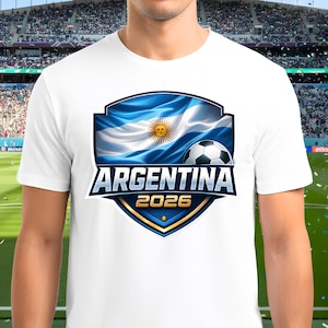 May include: White t-shirt featuring a shield design with the Argentina flag, a soccer ball, and the text "ARGENTINA 2026". The design is in blue, white, and gold colors. The shirt is worn by a person.
