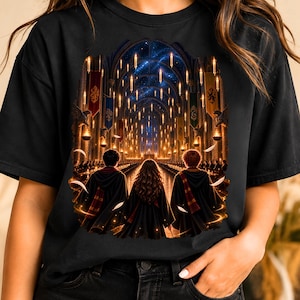 May include: A black t-shirt featuring a graphic print of three figures in dark robes standing in a grand hall with floating candles, banners, and a starry ceiling. The scene depicts a large interior space with long tables and architectural arches.