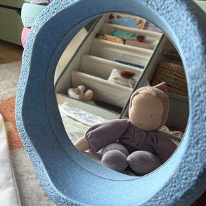 Puede incluir: Un espejo circular azul claro con una superficie texturizada. El espejo refleja una muñeca vestida con un atuendo morado. La muñeca está sentada frente a una estantería blanca con libros y otros objetos. El espejo es un objeto decorativo.