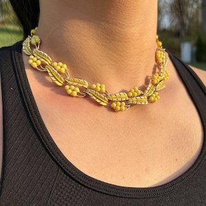 May include: A vintage necklace featuring a floral design with yellow beaded clusters and leaf accents. The necklace is made of gold-toned metal and is worn around the neck. The necklace is a statement piece.